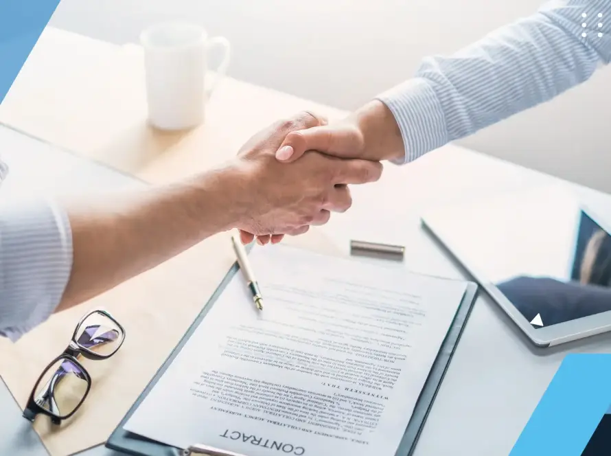 Dos personas se dan la mano sobre una mesa de oficina con papeles, unas gafas y una tablet.